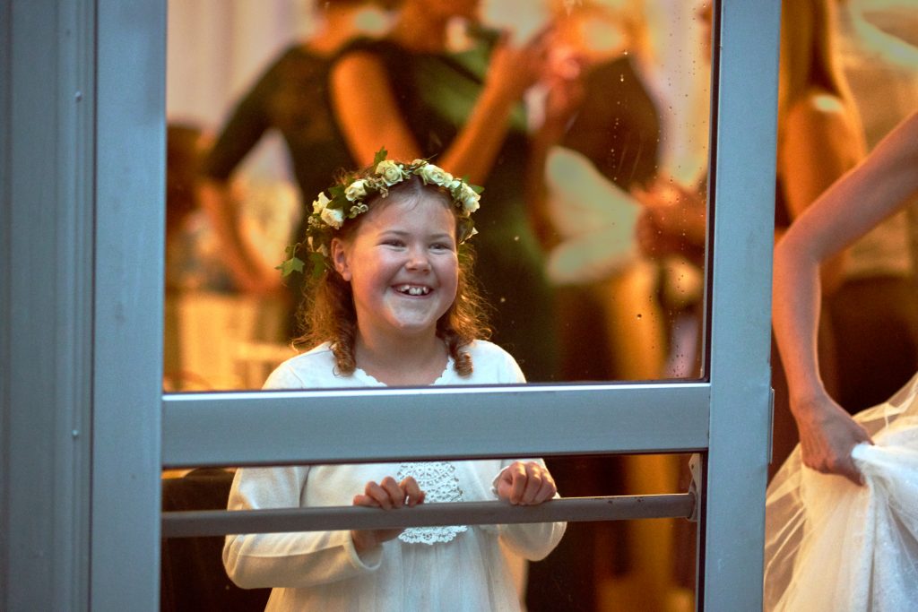 Young girl expectantly looking through a rainy window whist a sophisticated wedding occurs in the background