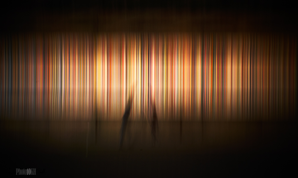 Blurred image of three passing pedestrians in front of the vertical line art under the bridge